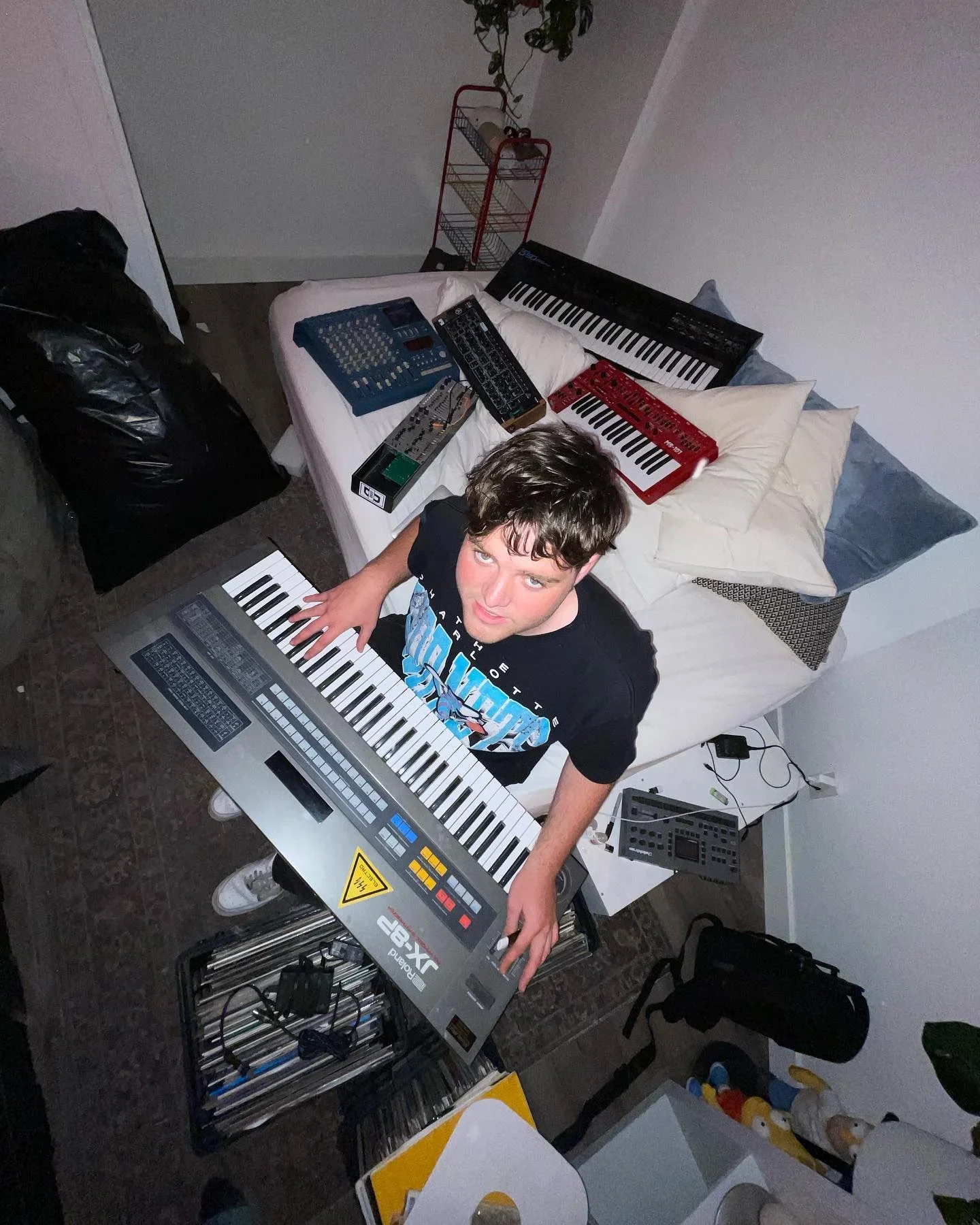 Cult Member looking up toward the camera, surrounded by synthesizers and music equipment.